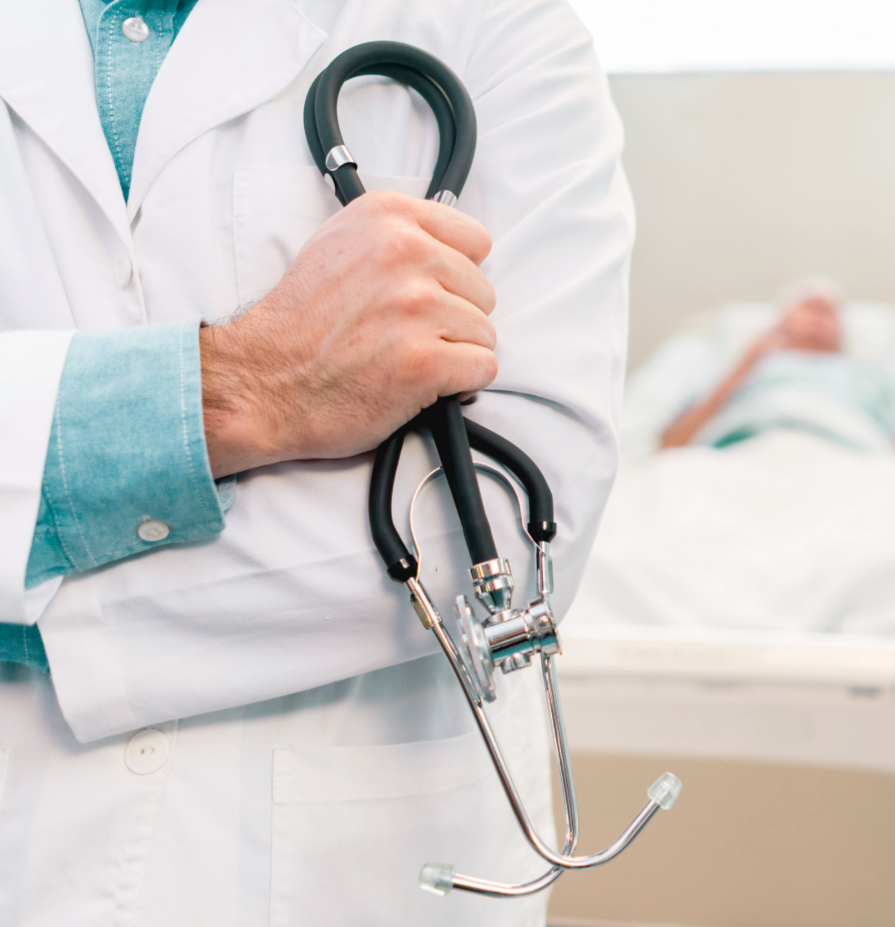 Close-up of a doctor wearing a white coat holding a stethoscope with a patient blurred on a hospital bed in the background.