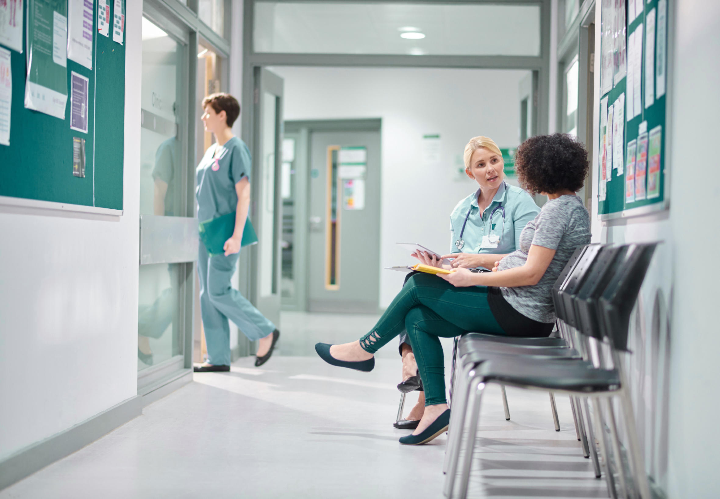 Female doctor talking with a pregnant woman seated in a clinic waiting area while another medical professional walks past.