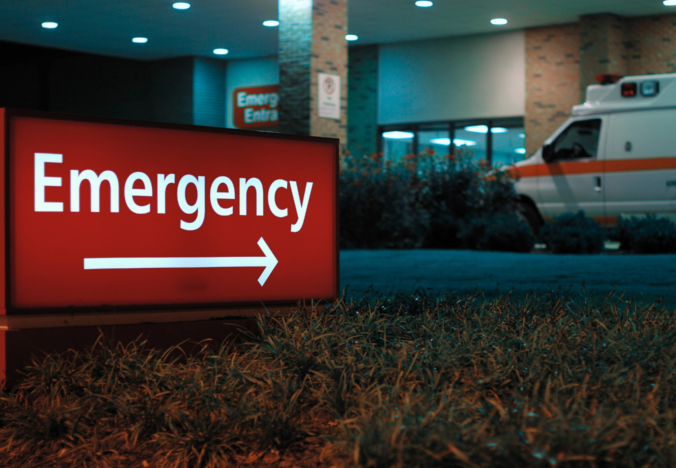 Illuminated red emergency sign with a right arrow outside a hospital at night, with an ambulance parked nearby.